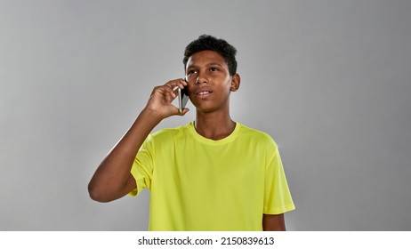 Thoughtful And Focused Sportive Black Boy Talking On Smartphone. Male Youngster Wearing Yellow T-shirt. Concept Of Modern Child Lifestyle. Isolated On Grey Background. Studio Shoot. Copy Space