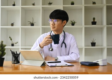 Thoughtful general practitioner in white uniform drinking coffee and looking through a window at medical office. - Shutterstock ID 2342250059