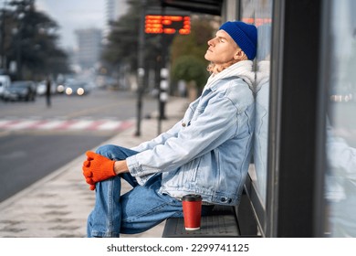 Thoughtful German hipster man sitting at bus stop with takeaway coffee waiting transport. Pensive Scandinavian guy looking distant clasping knee in gloves thinking about difficult life task solution - Powered by Shutterstock