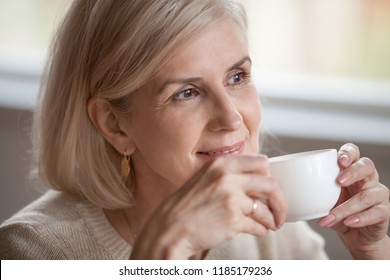 Thoughtful happy middle aged senior woman with beautiful face looking away drinking morning coffee, smiling mature old lady holding tea cup relaxing with positive thoughts dreaming enjoying wellbeing
