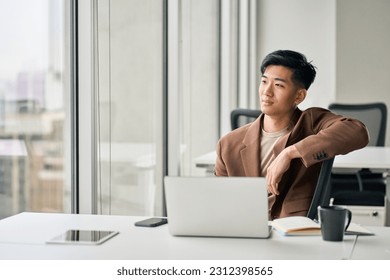 Thoughtful happy young Asian business man employee sitting with laptop in company office. Male professional manager executive looking at window thinking of future goals, dreaming or leadership.