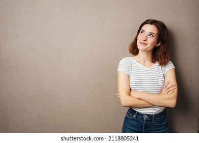 Thoughtful happy young woman smiling in anticipation as she stands with folded arms looking up to the side over a brown studio background with copyspace