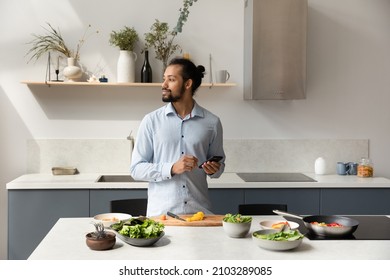 Thoughtful hipster guy consulting cooking online recipe on internet on mobile phone, thinking over dinner menu, looking away at table with vegetables, counter with fresh food ingredients - Powered by Shutterstock