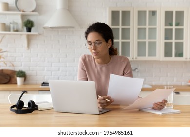 Thoughtful hispanic female sit at home workplace hold financial documents look on pc screen check data. Serious young woman think on business offer compare terms conditions in paper letters and online
