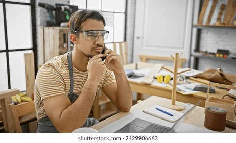 A thoughtful hispanic man with a beard wearing safety glasses sits in a carpentry workshop using a laptop and smartphone. - Powered by Shutterstock