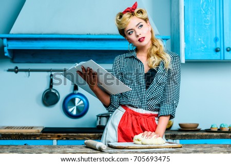 thoughtful housewife reading cookbook while making dough in kitchen