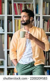 Thoughtful Indian Eastern Ethnic Man User Standing At Home In Front Of Bookcase Wearing Earbuds Drinking Coffee Thinking Looking Through Window Listening Music, Audio Book Or Podcast.