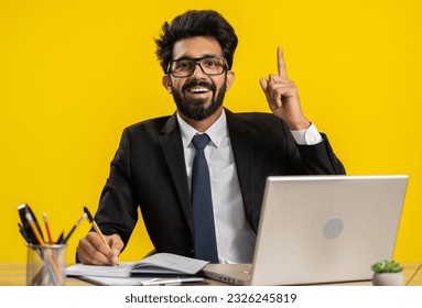 Thoughtful journalist indian businessman making notes, writing down thoughts with pen into notepad notebook diary, to do list, good idea at office workplace desk. Hindu man in suit working on laptop - Powered by Shutterstock