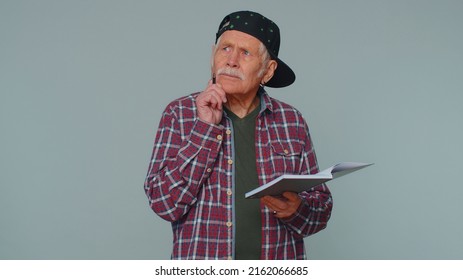 Thoughtful Journalist Senior Man Making Notes, Writing Down Thoughts With Pen Into Notepad Notebook Diary, To Do List, Good Idea. Elderly Grandfather Isolated Alone On Gray Studio Wall Background