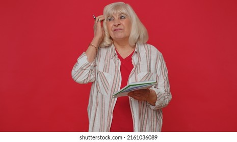 Thoughtful Journalist Senior Woman Making Notes, Writing Down Thoughts With Pen Into Notepad Notebook Diary, To Do List, Good Idea. Elderly Grandmother Isolated Alone On Red Studio Wall Background