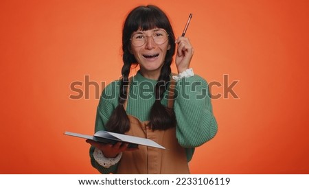 Thoughtful journalist woman with in green sweater making notes, writing down thoughts with pen into notepad notebook diary, to do list, good idea. Young adult girl isolated on orange studio background