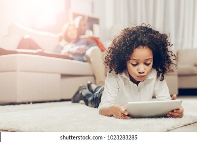 Thoughtful kid looking at tablet while resting on soft tapis inside. Focus on girl. Parents on background