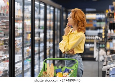 Thoughtful lady with shopping cart looking at fridge with daily products in grocery store and choosing what to buy. Woman buying groceries in supermarket. - Powered by Shutterstock