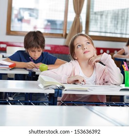 Thoughtful little schoolgirl looking up while using digital tablet with classmates in background at classroom - Powered by Shutterstock