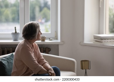 Thoughtful lonely retired woman sitting on couch at home, looking at window away in deep thoughts, thinking over health problems, bad news, loss, suffering from depression, making hard decision