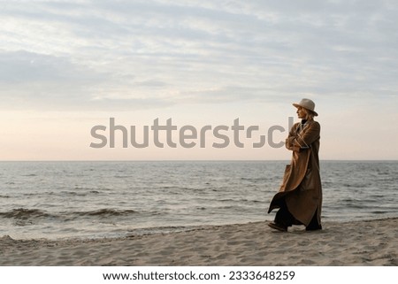 Thoughtful lonely stylish senior woman in brown coat and hat walking on beach in evening, copy space. Adult caucasian lady alone looking at sea.