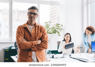 Thoughtful male employee in casual clothes and glasses standing against colleagues working on project with crossed arms and looking away - Shutterstock ID 2340604493