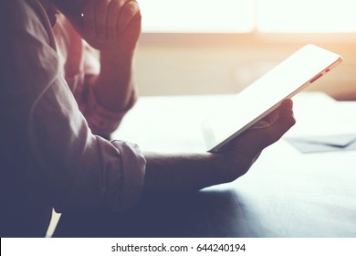 Thoughtful male person looking to the digital tablet screen while sitting in modern loft interior at the table, experienced entrepreneur reading some text or electronic book at the office, filter sun