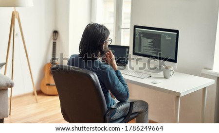 Thoughtful male web developer sitting at his workplace, looking at screen with programming code, working from home. Using desktop computer and laptop. Freelance, home office. Stay home, self isolation