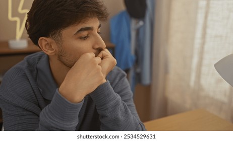 A thoughtful man with a beard in a casual blue sweater sits pensively in a cozy room at home. - Powered by Shutterstock