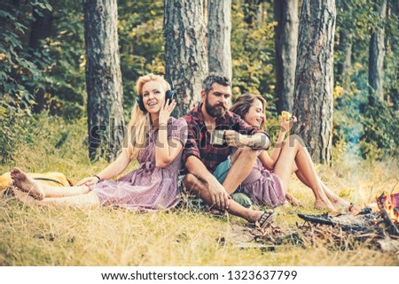Thoughtful man with beard drinking tea of coffee while watching flames of campfire. Blond girl listening to music while her sister is eating apple. Various perception of perfect relaxation.