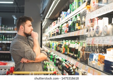 Thoughtful man with a beard looks at bottles with beer in a supermarket. Man wears a shirt, looks at the shelf with bottles of beer and thinks. Buyer buys a beer at a supermarket. - Powered by Shutterstock