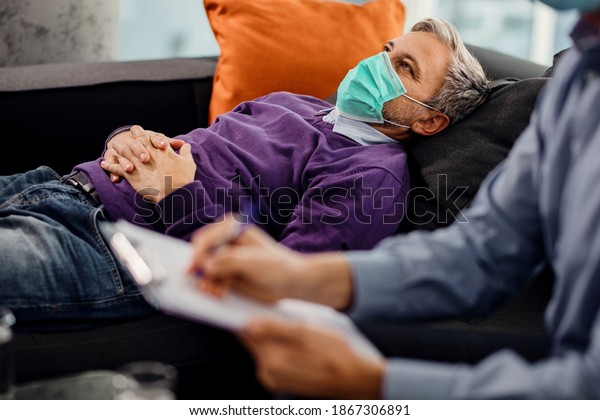 Thoughtful man with face mask lying down on\
psychiatrist\'s couch during a therapy session with mental health\
professional. 