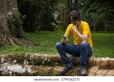 Thoughtful man looking at smartphone while sitting on natural log outdoors	