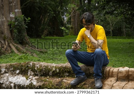 Thoughtful man looking at smartphone while sitting on natural log outdoors	