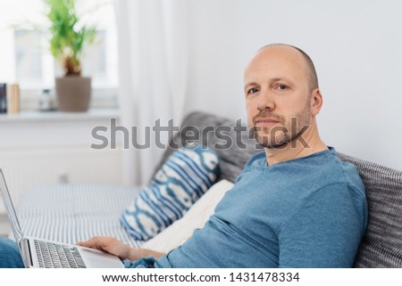 Thoughtful man scrutinising the camera as he sits relaxing on a couch at home with his laptop computer