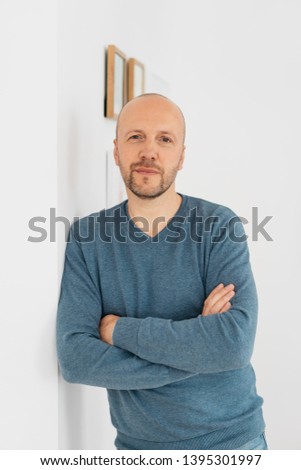 Thoughtful man scrutinising the camera with an intent stare and folded arms as he leans against a white interior wall