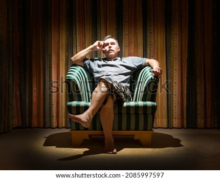 Thoughtful man is sitting in a dark room in a vintage armchair under the light of a lamp