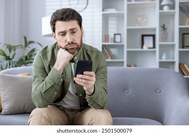 Thoughtful man sitting on sofa looking at smartphone at home. Pensive expression, appears deep in thought or concerned. Casual indoor setting with focus on man's face and phone. - Powered by Shutterstock