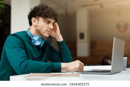 Thoughtful man student with headphones around his neck is studying or working on laptop, with hand to his temple, in bright office or classroom space - Powered by Shutterstock