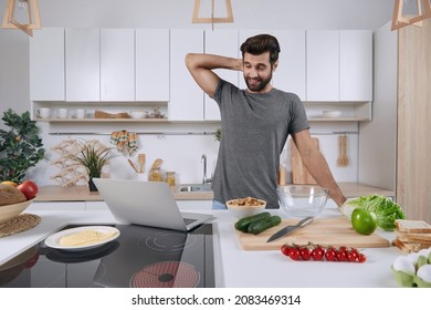 Thoughtful Man Watching Something On Laptop Computer While Cooking At Home Kitchen. Concept Of Dieting, Vegetarian And Healthy Eating. Young Caucasian Guy Standing At Table With Organic Products