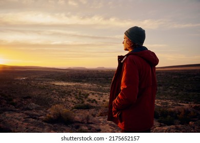 Thoughtful man wearing jacket and cap standing on mountain looking at morning sunrise