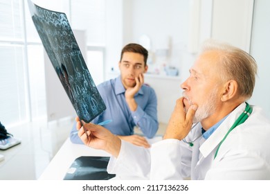 Thoughtful Mature Adult Male Physician Consult Frustrated Young Man Patient Giving Bad News Explaining Results Of MRI Image. Unhappy Scared Young Man Listening To Bad News Sitting In Doctor Office.