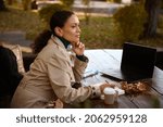 Thoughtful mature African woman rests in outdoor cafe, sits at a wooden table with fallen dry oak leaves with takeaway craft paper cup of hot drink, makes notices on her notepad and works on laptop