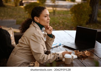 Thoughtful mature African woman rests in outdoor cafe, sits at a wooden table with fallen dry oak leaves with takeaway craft paper cup of hot drink, makes notices on her notepad and works on laptop