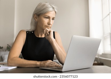 Thoughtful mature freelancer woman working at laptop at home, looking at screen with thoughtful face, touching chin, thinking, reading, watching online content, making decision in deep thoughts - Powered by Shutterstock