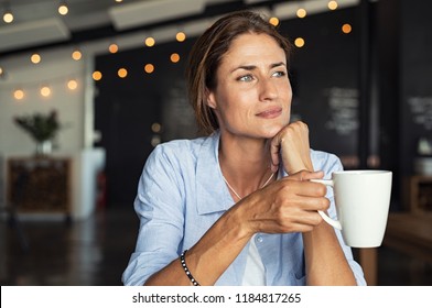 Thoughtful mature woman sitting in cafeteria holding coffee mug while looking away. Middle aged woman drinking tea while thinking. Relaxing and thinking while drinking coffee. - Shutterstock ID 1184817265