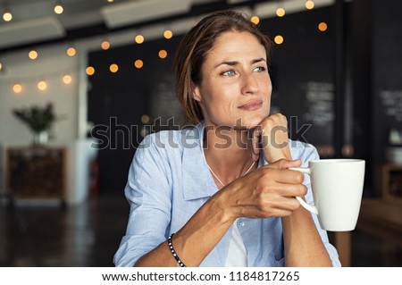Thoughtful mature woman sitting in cafeteria holding coffee mug while looking away. Middle aged woman drinking tea while thinking. Relaxing and thinking while drinking coffee.