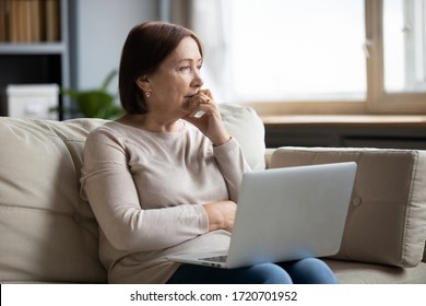 Thoughtful Mature Woman Using Laptop, Sitting On Couch At Home Alone, Pensive Older Female Thinking About Finance Or Health Problem, Looking In Distance, Having Doubts, Feeling Lonely