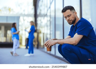 A thoughtful medical professional in blue scrubs sits outdoors holding a coffee cup. In the background, two colleagues engage in conversation, highlighting teamwork and healthcare collaboration. - Powered by Shutterstock