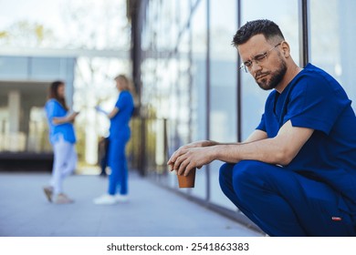 A thoughtful medical professional in blue scrubs holding a coffee cup, reflecting during a break outside a hospital building. Colleagues are seen in the background, engaging in conversation. - Powered by Shutterstock