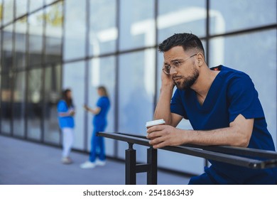 A thoughtful medical professional in blue scrubs leans against a railing while holding a coffee cup. Two colleagues are seen conversing in the background outside a modern building. - Powered by Shutterstock