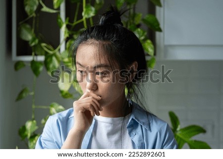 Thoughtful melancholic Asian woman touches lips with hand experiencing psychological disorder. Unhappy Chinese female stands in room near closet with houseplants and needs help of psychotherapist.