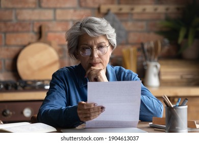 Thoughtful Middle Aged 60s Woman In Eyeglasses Reading Paper Letter Carefully. Pensive Focused Older Mature Retired Lady Looking At Correspondence Sheet. Considering Bank Notification At Home.