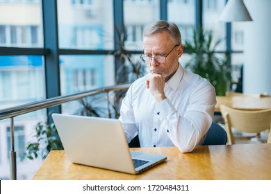 Achtsamer Geschäftsmann mittleren Alters in weißem Hemd. Mann sitzt mit einem Laptop in der Nähe des Fensters arbeitet. Geschäftskonzept.