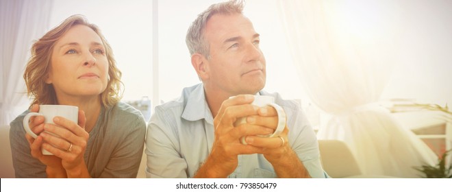 Thoughtful middle aged couple sitting on the couch having coffee looking away at home in the living room